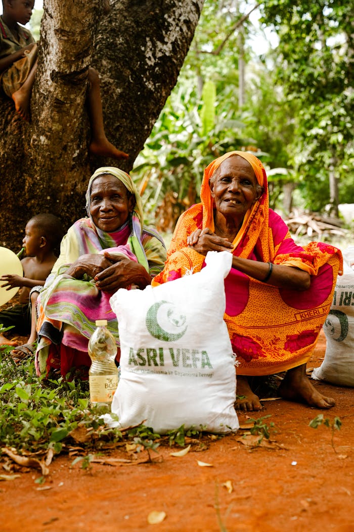 about-01 Two elderly women hold aid packages in a rural outdoor setting, sitting on red soil.