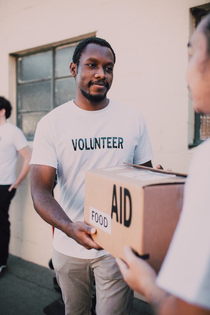 hero-img-01 A volunteer hands a box labeled 'aid' and 'food' during an outdoor charity event.