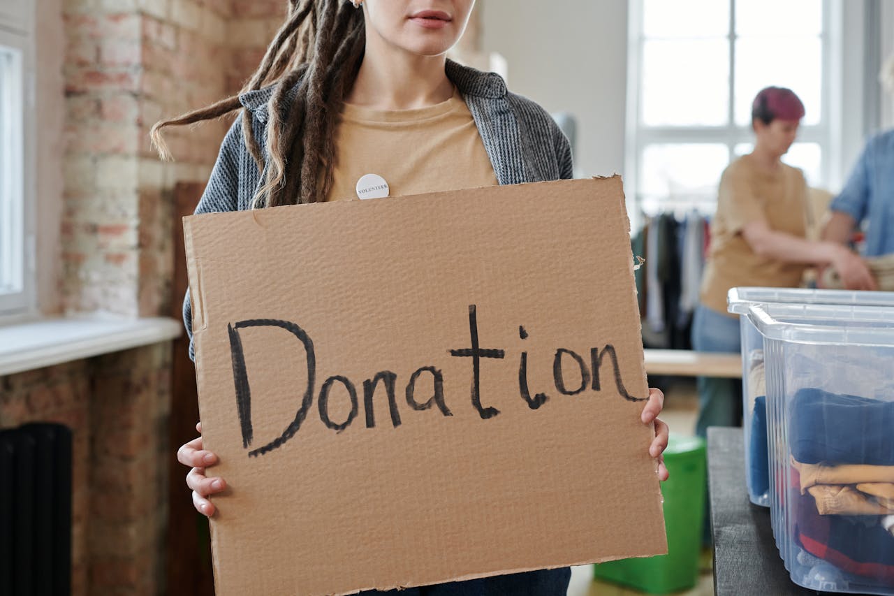gallery-2 Volunteer holding a cardboard donation sign in a community center with clothing and supplies.