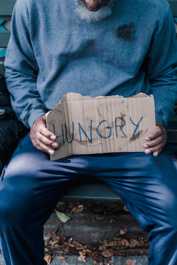 gallery-4 Man seated on bench holding cardboard sign with 'HUNGRY' text, highlighting urban poverty.